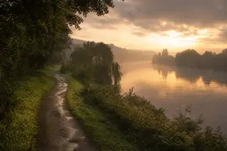 Chemin de halage verdoyant bordant la Seine à l’aube, lumière dorée, atmosphère humide et paisible, perspective douce sur la rive.